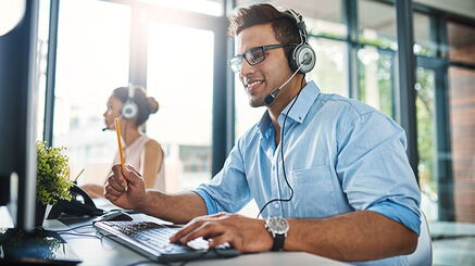 A young man with a headset is sitting at a desk in front of a screen, one hand on the keyboard. A young woman, also wearing a headset, can be seen in the background.